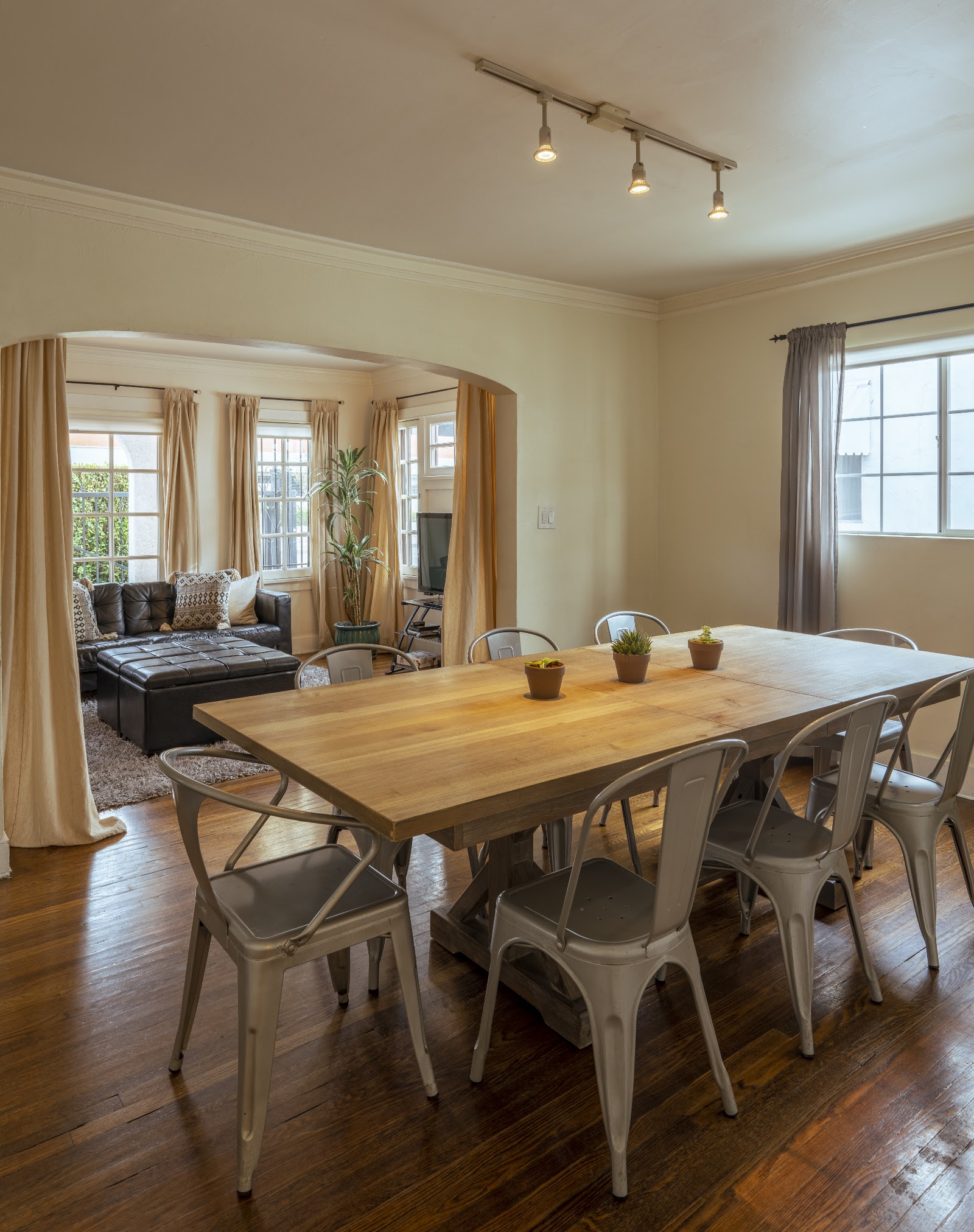 Interior dining space with wooden table, white chairs, large windows with curtains, and adjacent lounge seating area