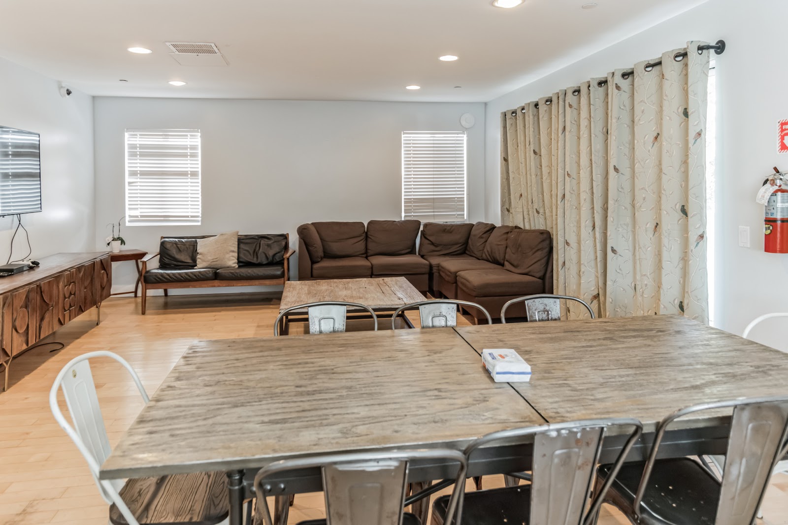 Interior common room with large dining table, sectional sofa, television, windows with curtains, and wooden flooring