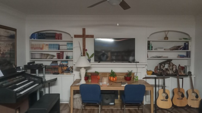 Interior recreation room with piano, guitars, desk seating area, bookshelf, mounted television, and wooden cross decoration