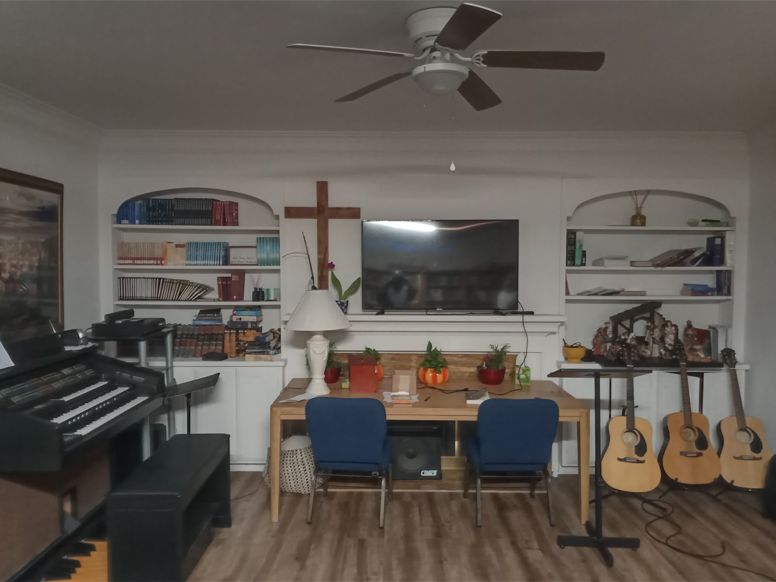Interior recreation room with piano, guitars, desk seating area, bookshelf, mounted television, and wooden cross decoration