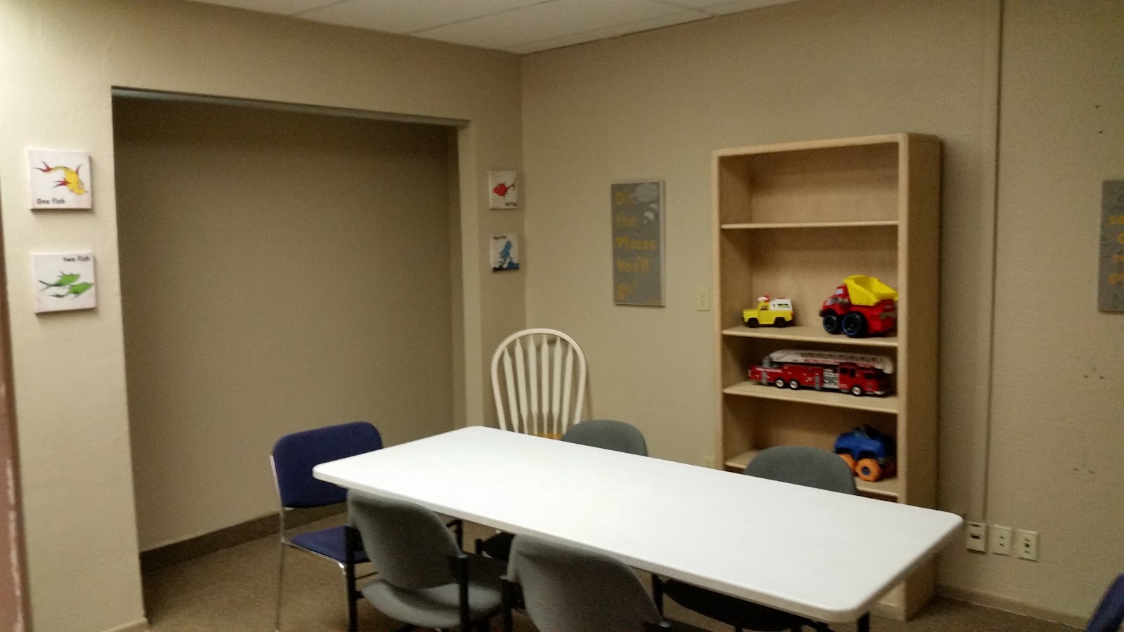 Interior room with white rectangular table, multiple gray chairs, wooden shelving with toys and materials.