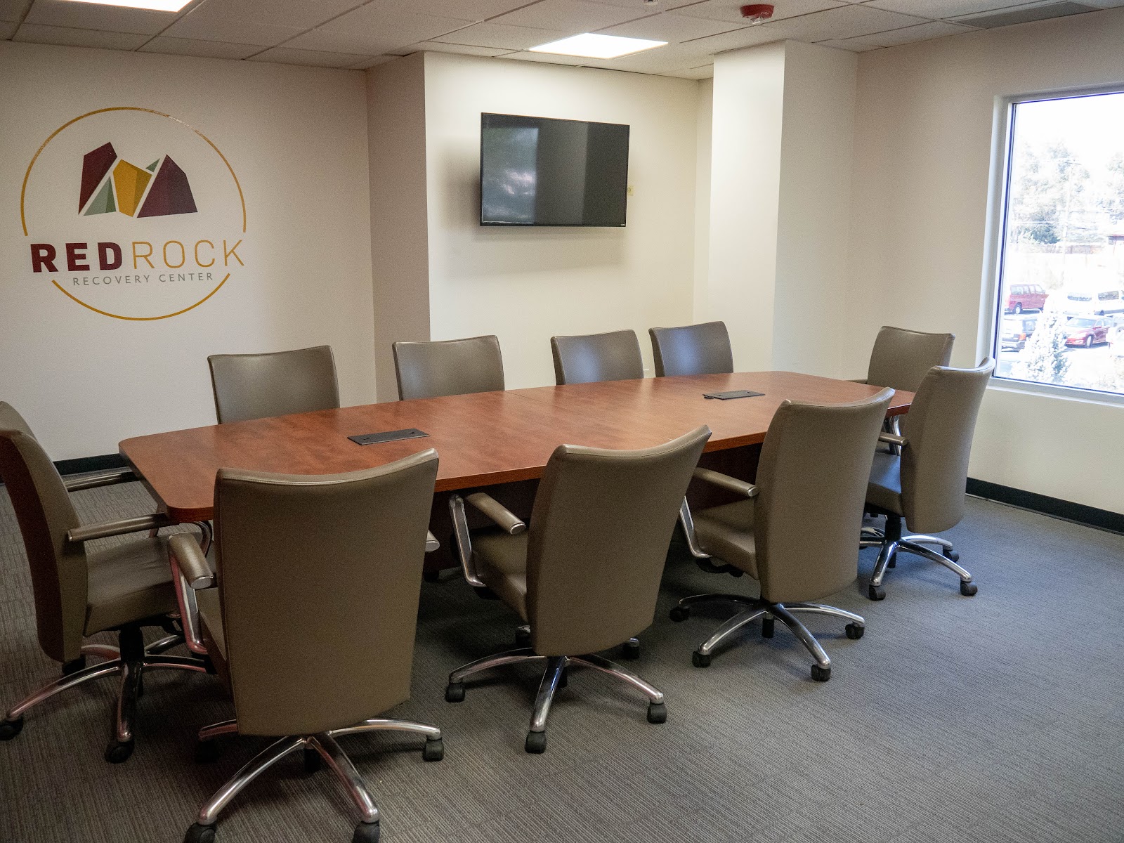 Interior conference room with large wooden table, executive chairs, mounted television, and windows with outside views