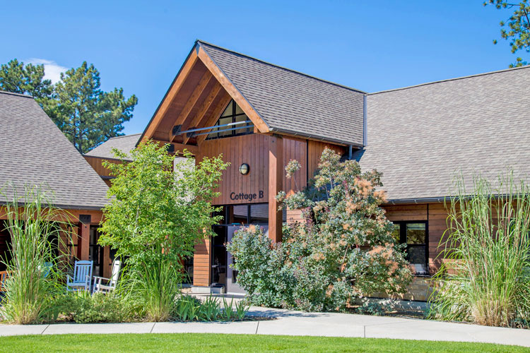 Exterior cottage building with wood siding, gable entrance, landscaped grounds with shrubs and ornamental plantings.