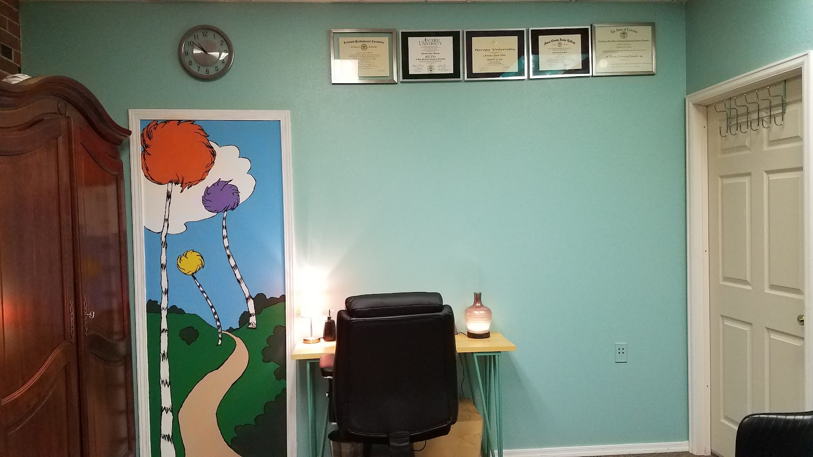 Interior office space with turquoise walls, framed professional certificates, colorful artwork, black chair, and small side table with lamp