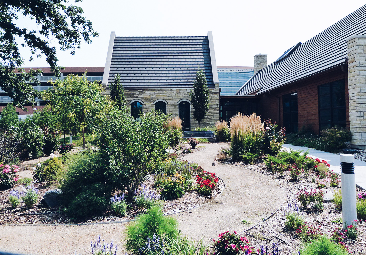 Outdoor courtyard area with multiple modern buildings, stone construction, landscaped gardens with flowering plants and paved walkways.