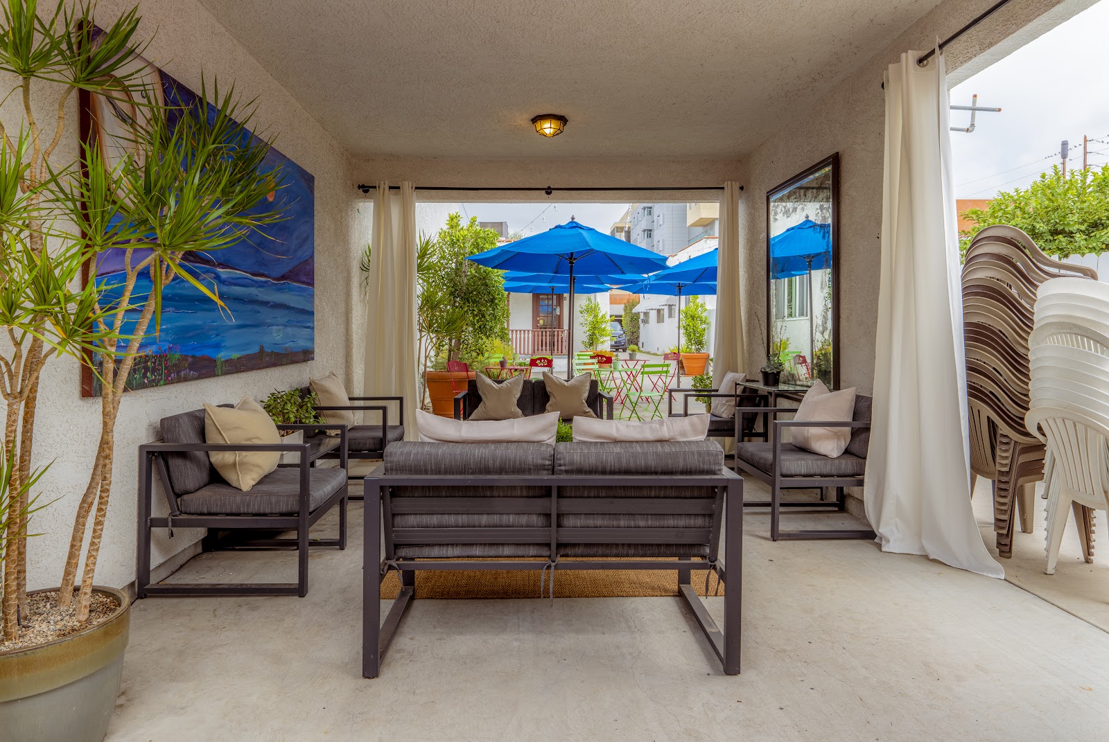 Interior covered patio with modern seating, blue umbrella visible outside, large windows, potted plants, and wall art
