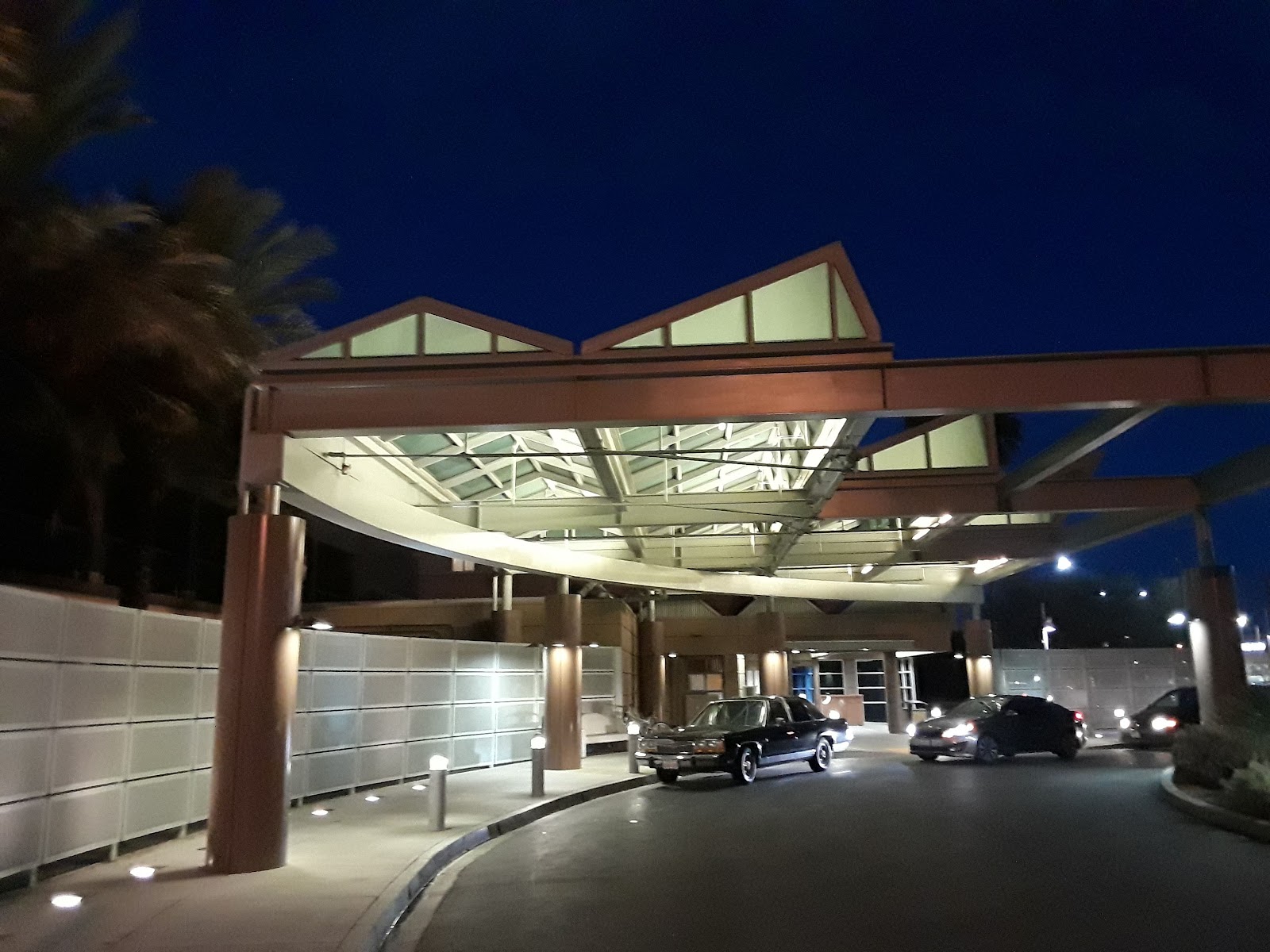 Exterior emergency entrance with illuminated metal canopy structure, glass doors, and vehicles in covered drive-through area