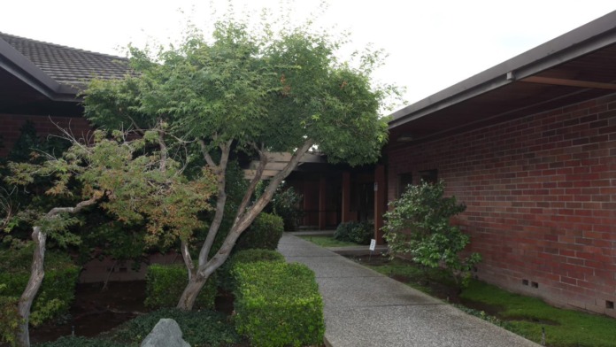 Exterior showing landscaped grounds with mature trees, gravel pathway, rock features, and brick building facade