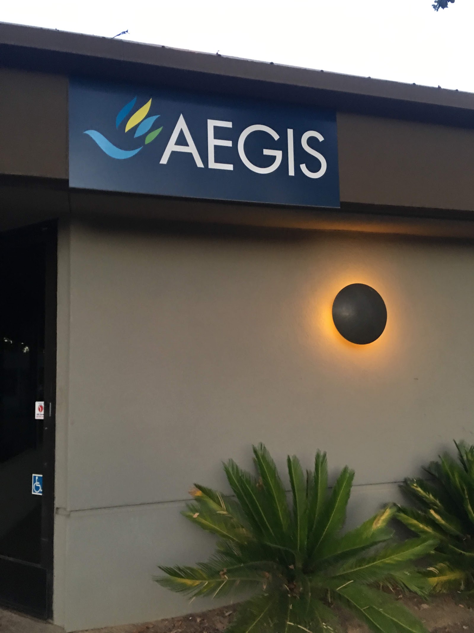 Exterior entrance showing Aegis Treatment Centers blue and white logo sign mounted above entrance with ground-level landscaping