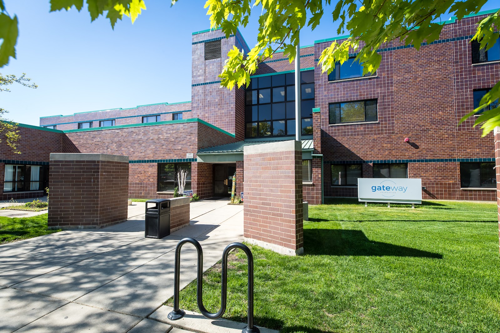 Exterior brick building with gateway sign, bicycle racks, manicured lawn, and modern architectural design with turquoise accents