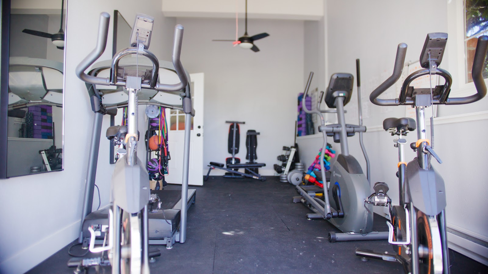 Interior fitness center with multiple stationary bikes lined against walls, black flooring, ceiling fan, and natural lighting.