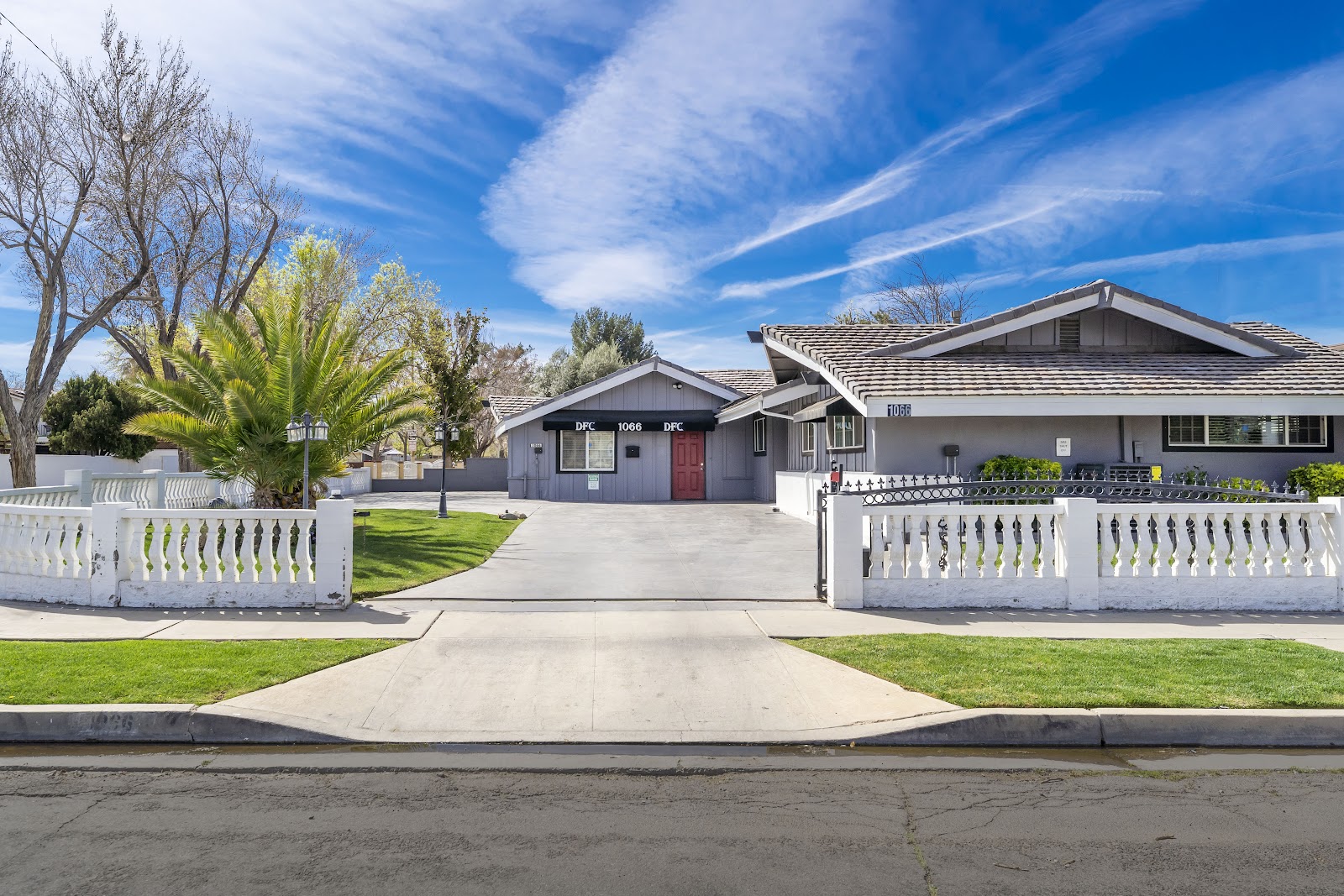 Exterior showing facility driveway approach, white balustrade fencing, landscaped grounds with palm and deciduous trees, and multi-building complex.