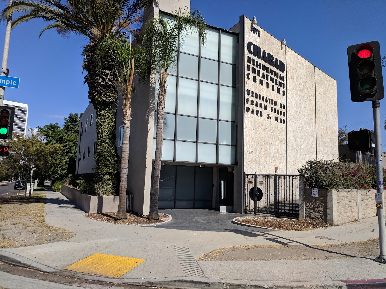 Exterior street view of institutional building with modern facade, large windows, palm tree landscaping, and entrance approach