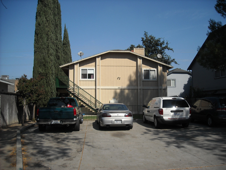 Exterior view of tan modular home with entry stairs, driveway with parked vehicles, cypress trees, and neighboring residential buildings