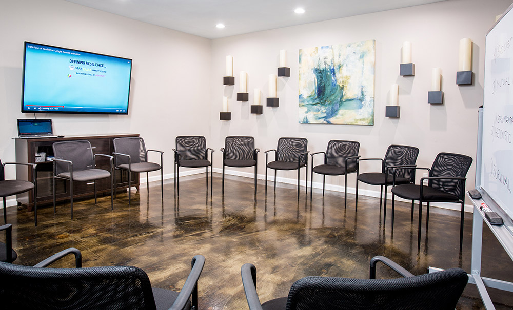 Interior circular meeting space with office chairs arranged in circle, flat-screen TV, whiteboard, wall art, and polished flooring