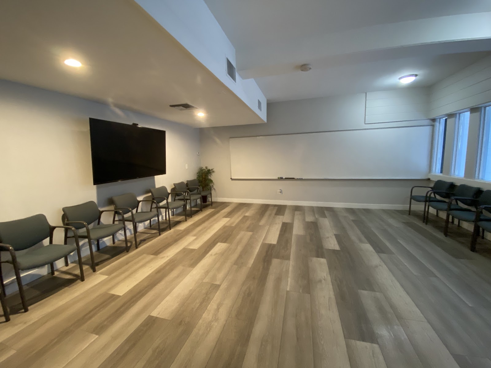 Interior meeting space with wooden flooring, rows of gray chairs, wall-mounted TV, whiteboard, and windows