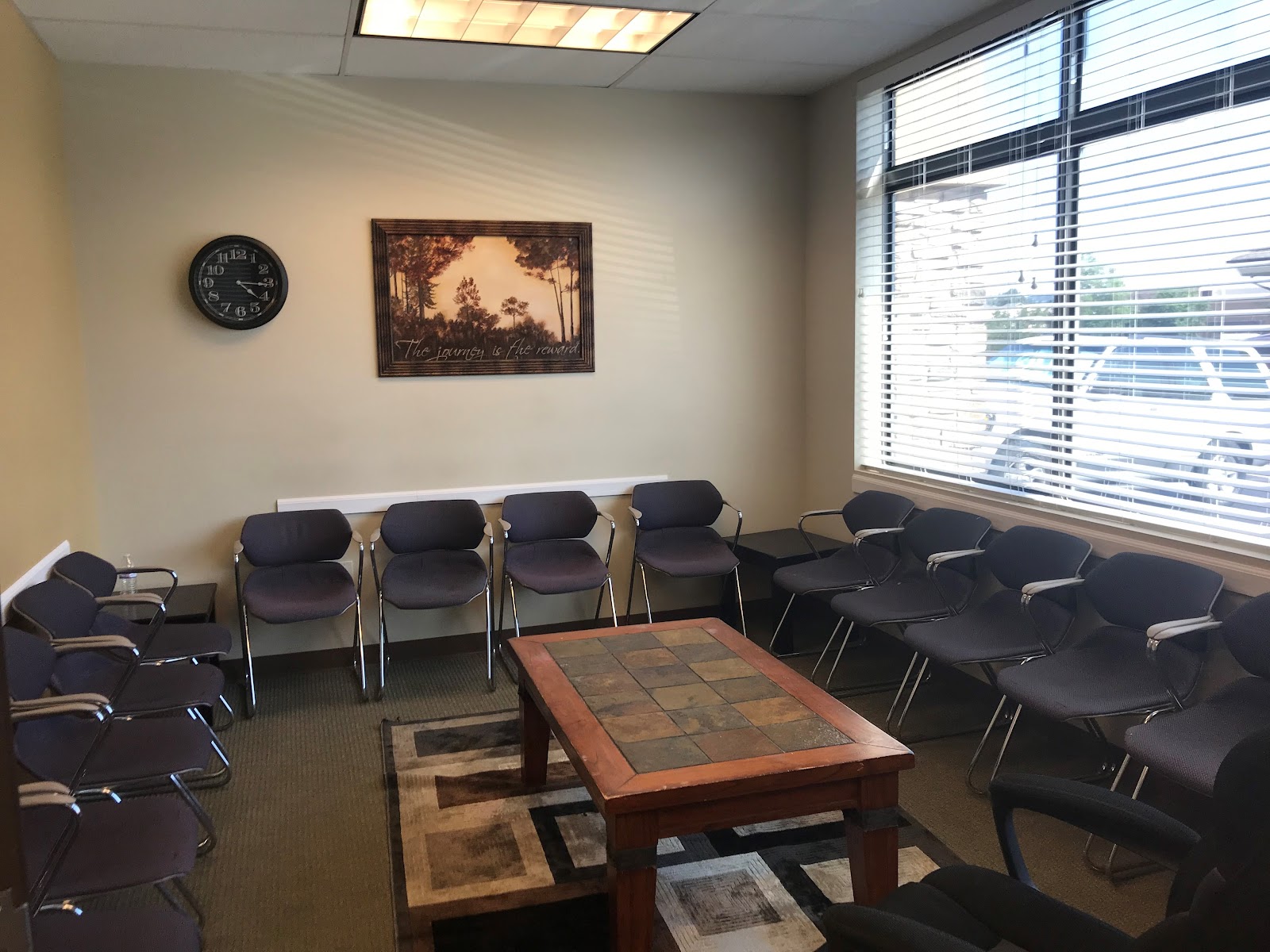 Interior therapy room with multiple black chairs arranged in circle, wooden table, wall clock, framed art, and window with blinds