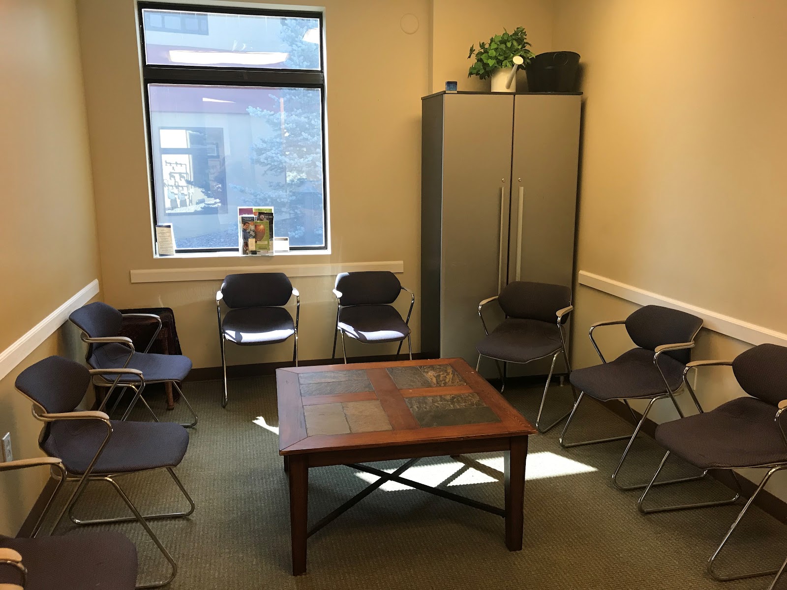 Interior waiting room with multiple black chairs arranged around wooden table, yellow walls, large windows, and decorative wall art