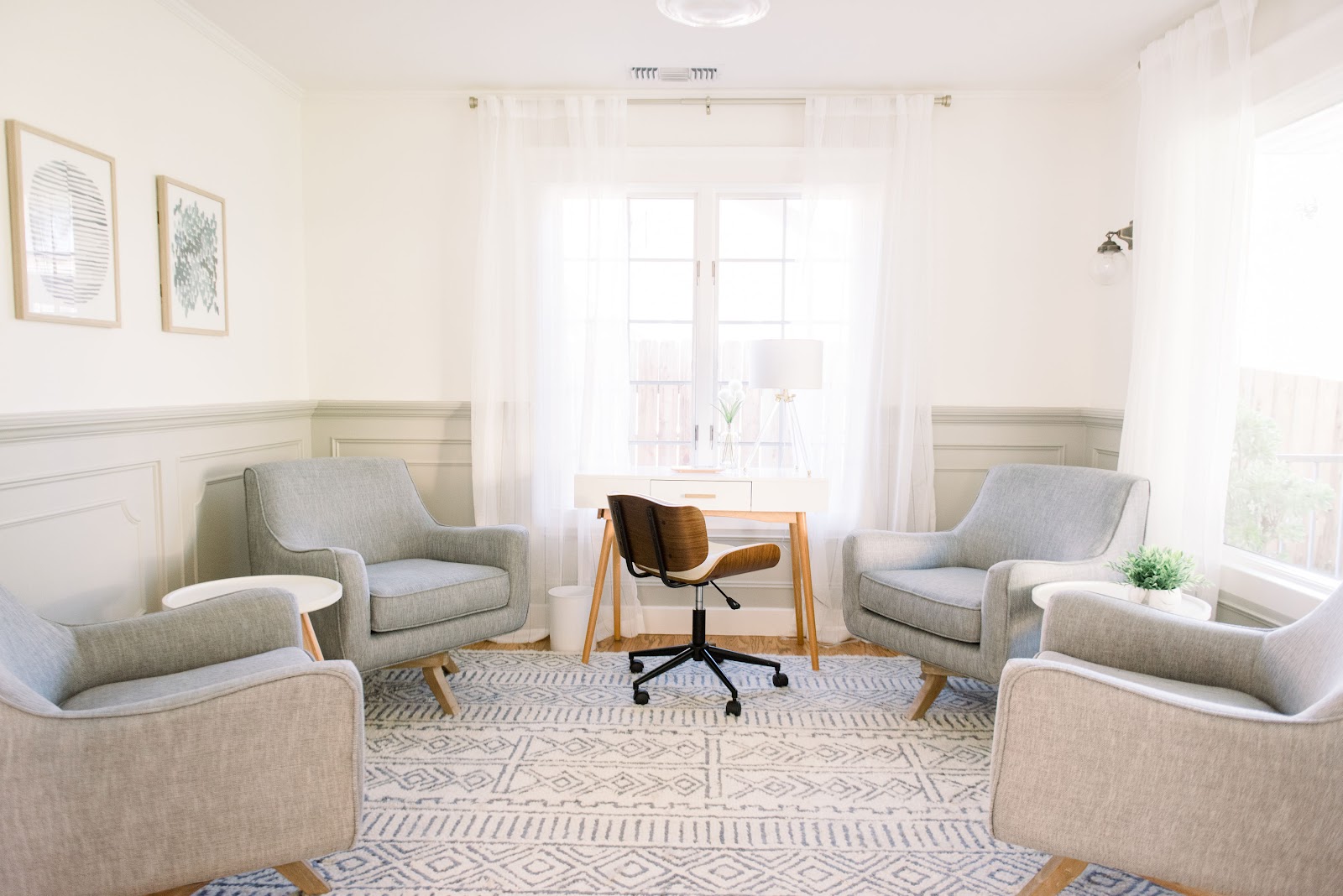 Interior therapy room with four upholstered armchairs, wooden desk, large windows with white shutters, and calm neutral decor
