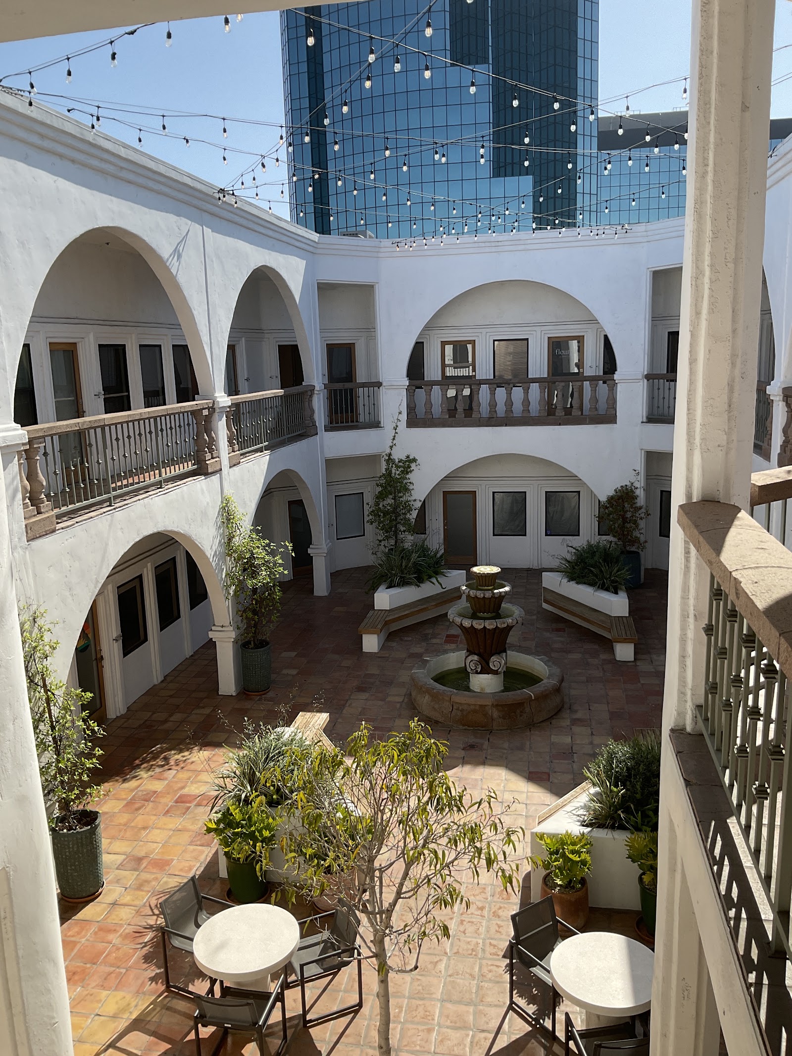 Three-story interior courtyard with white arched architecture, terracotta flooring, central fountain, potted plants, and modern glass ceiling