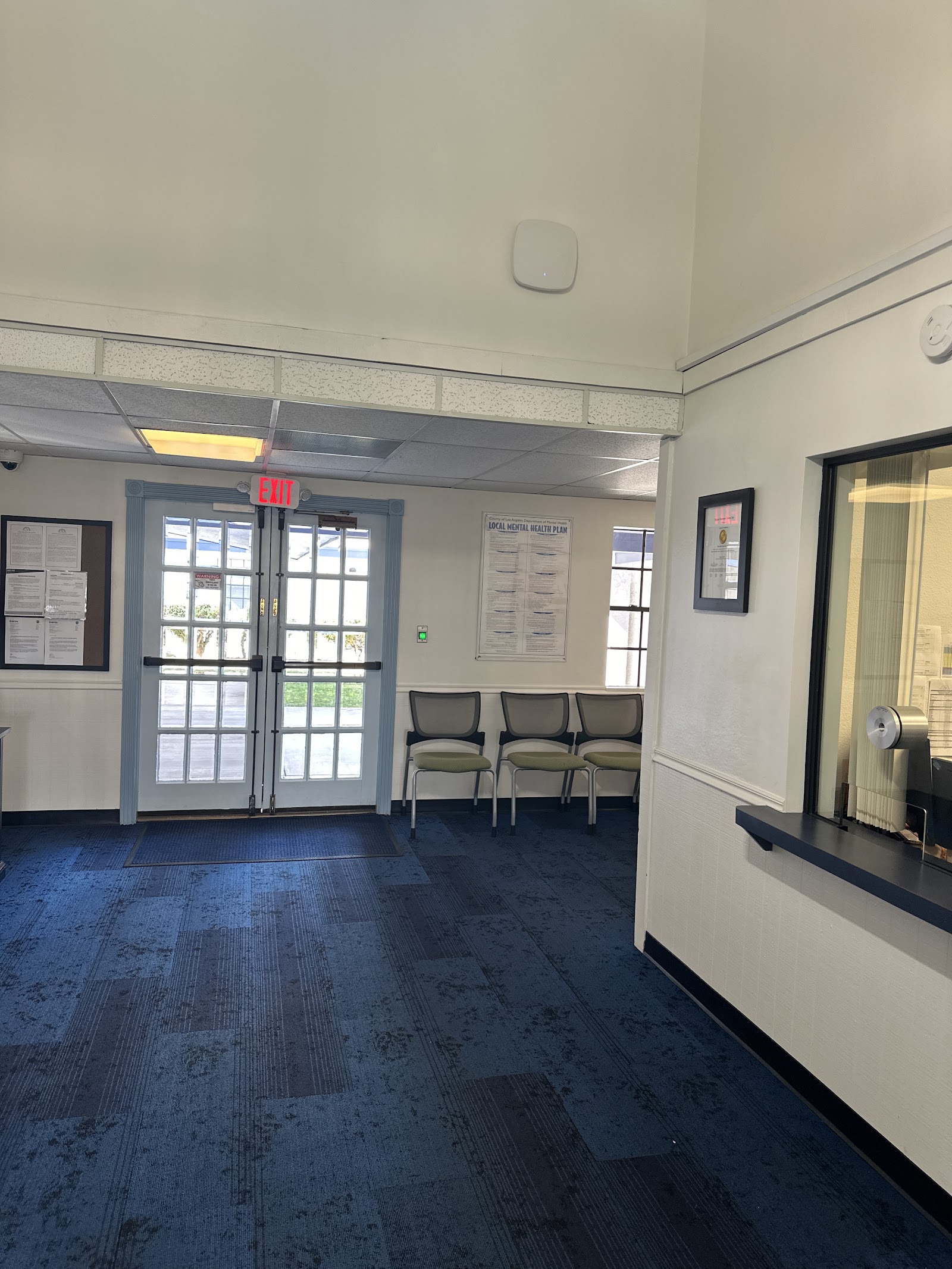 Interior lobby with glass doors featuring exit sign, seating area with gray chairs, information boards, and carpeted flooring