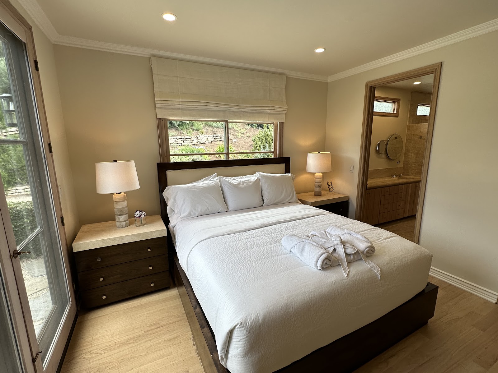 Interior patient bedroom with queen bed, dark wood furniture, ensuite bathroom, and glass door to outdoor terrace with landscaping