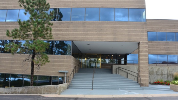 Modern facility entrance with horizontal tan siding, large glass windows, metal railings, concrete steps, and landscaping