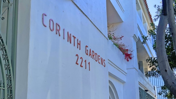 Exterior of white building with red painted address text reading Corinth Gardens 2211 and decorative metal gate