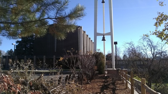 Exterior of modern dark brick treatment facility with tall bell tower structure, landscaped grounds, and surrounding evergreen trees.