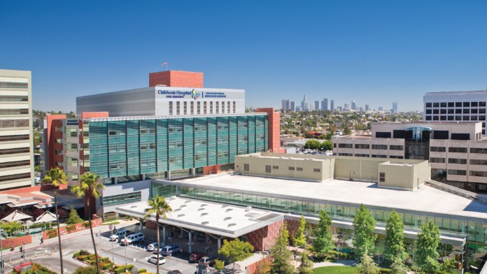 Exterior view of Children's Hospital Los Angeles Adolescent Medicine facility showing main building with turquoise glass panels, signage, parking area, and Los Angeles skyline in background