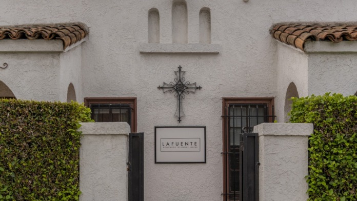 Exterior front entrance of Spanish colonial-style treatment center with white stucco, ivy-covered pillars, decorative cross, and LAFUENTE signage