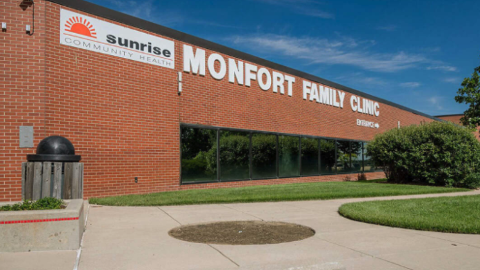 Exterior brick building with Sunrise Community Health and Monfort Family Clinic signage, landscaped grounds, and parking area