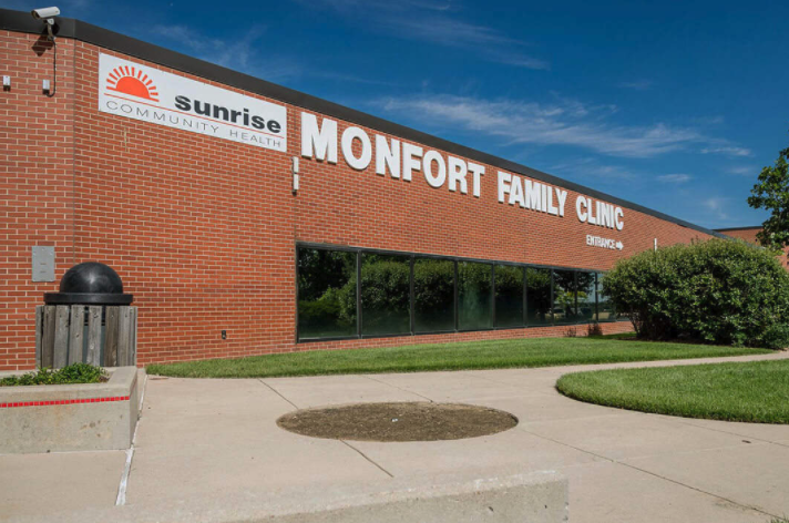 Exterior brick building with Sunrise Community Health and Monfort Family Clinic signage, landscaped grounds, and parking area