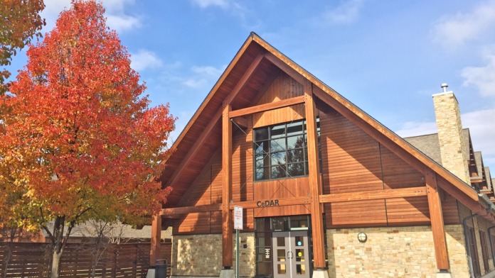 Exterior of treatment facility showing modern wood and stone entrance with gabled roof, landscaping, and blue sky backdrop.