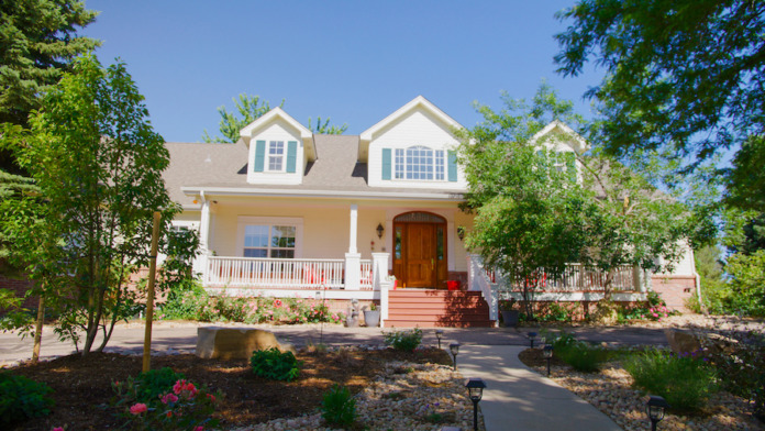 Exterior two-story house with cream and white siding, dormers, red brick chimney, front porch with white columns, and landscaped grounds.