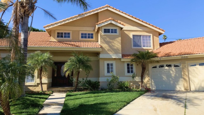 Exterior view of tan two-story residential building with terra cotta tile roof, palm trees, manicured lawn, and driveway