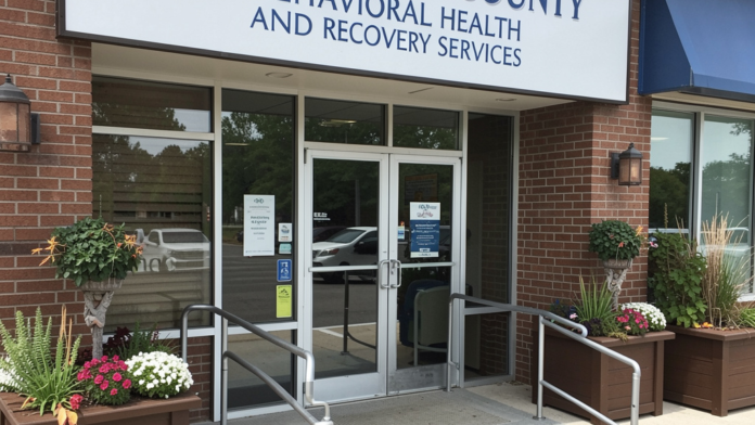 Exterior front entrance of brick treatment facility with blue facility signage, glass doors, and landscaped planters with flowering plants.