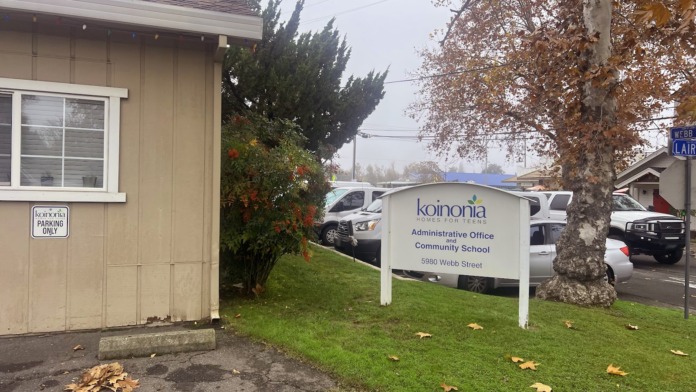 Exterior of Koinonia Family Services facility showing tan building, white entrance sign, landscaping, and parked vehicles