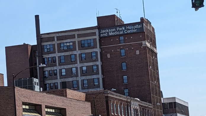 Exterior view of multi-story brick hospital building with Jackson Park Hospital and Medical Center signage, street view with parked vehicles