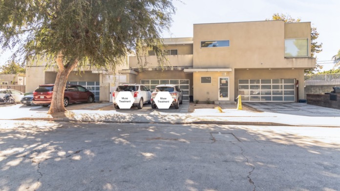 Exterior front view of Muse Treatment facility showing tan-colored building with modern architecture, glass doors, parking area, and branded vehicles