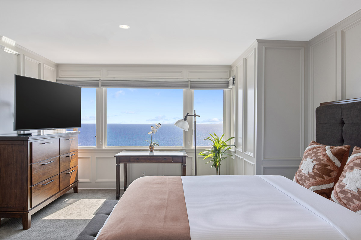 Interior master bedroom with large bed, ocean views through windows, desk, fireplace, and elegant furnishings