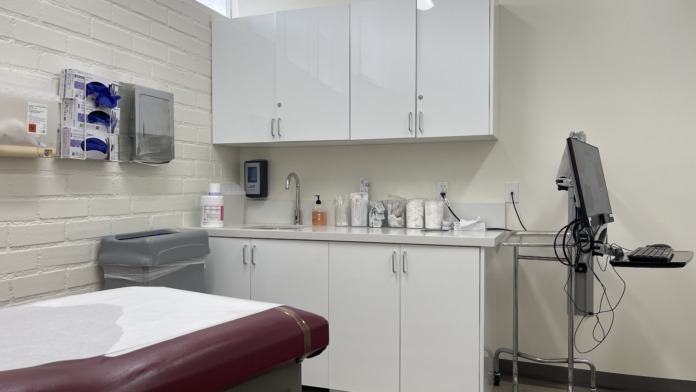 Interior clinical exam room with medical cabinets, sink, treatment table, and wall-mounted equipment