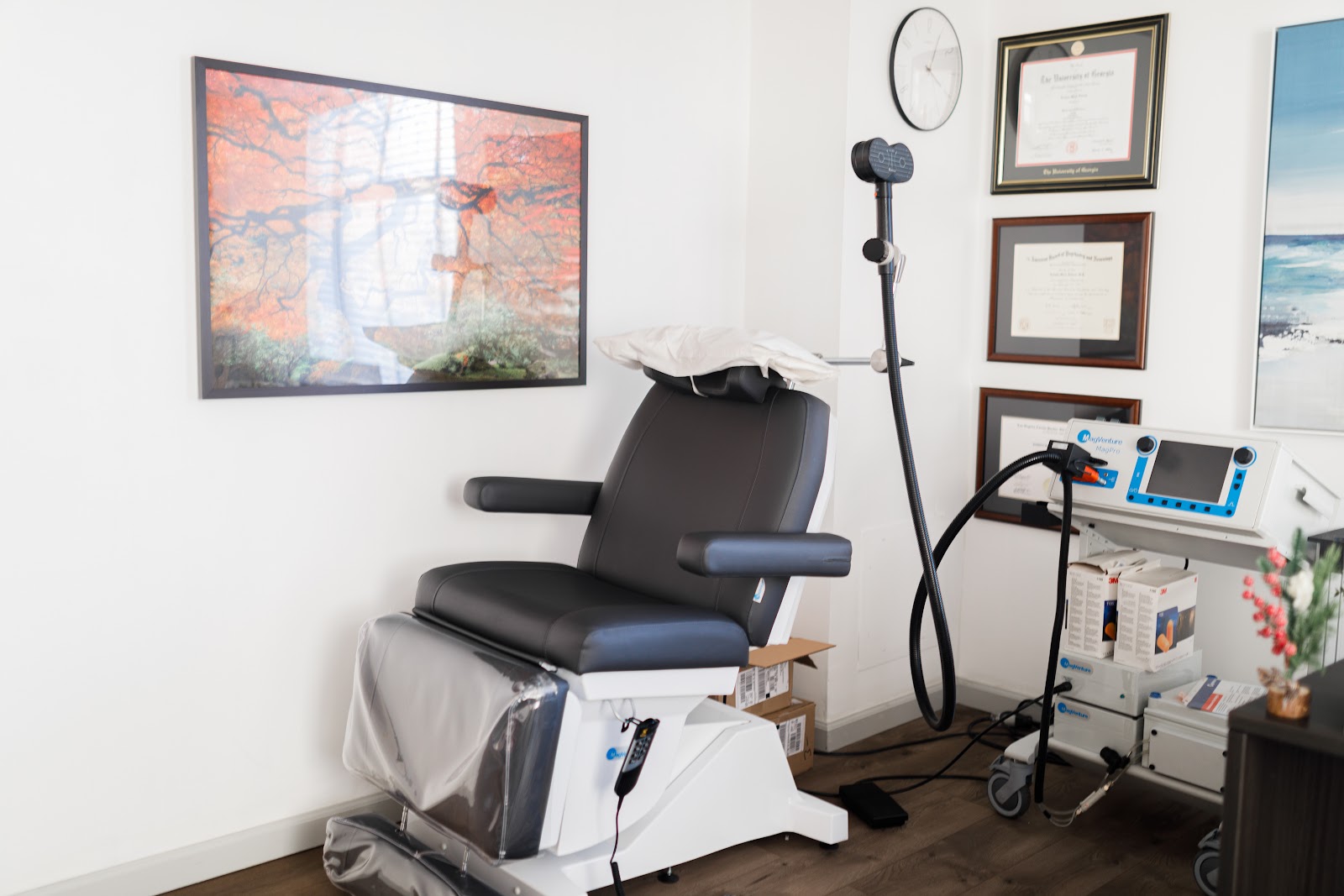 Interior treatment room with reclined patient chair, medical equipment cart, wall clock, and framed landscape artwork