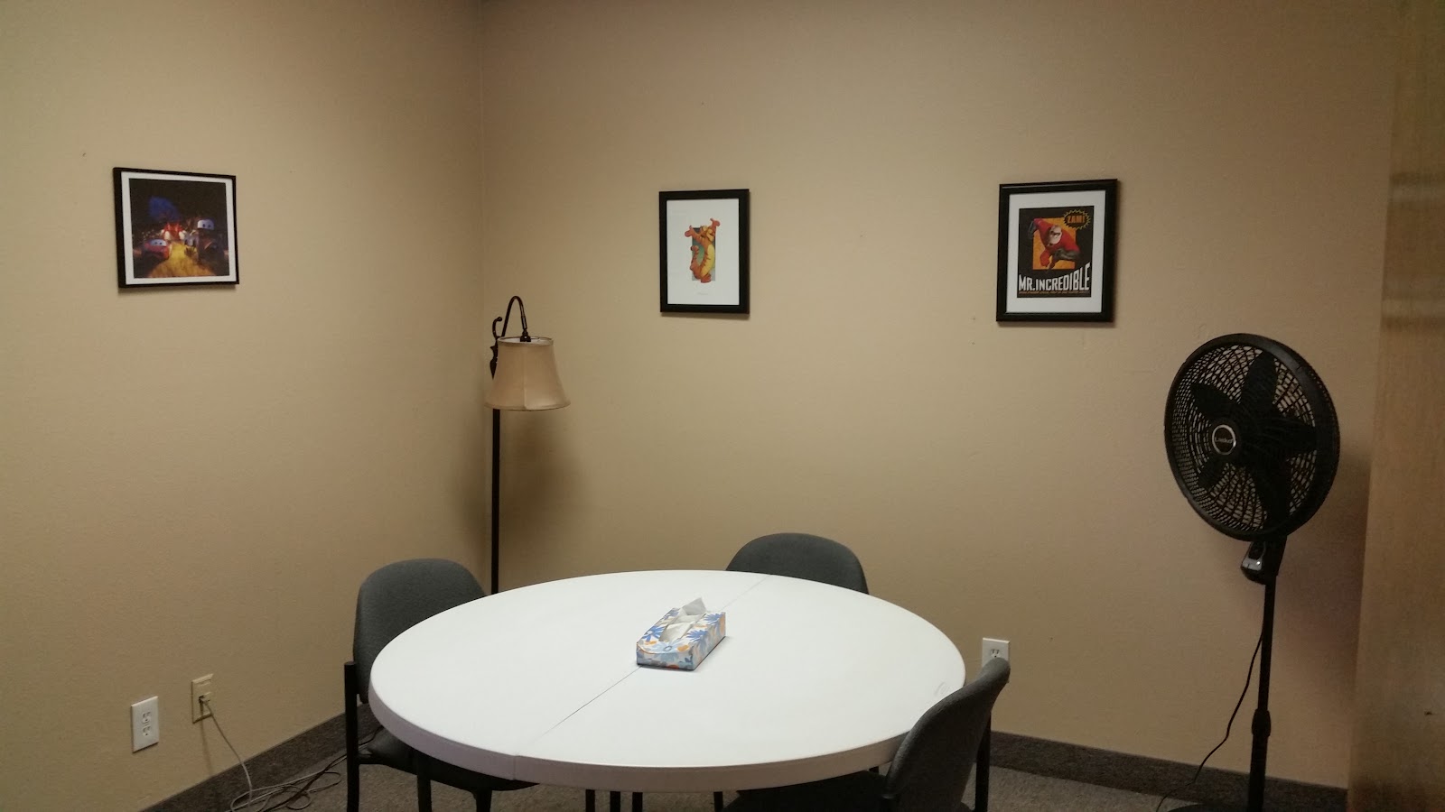 Interior room with white round table, gray chairs, framed artwork, floor lamp, and standing fan.
