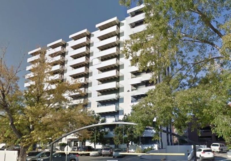 Exterior view of modern multi-story white institutional building with multiple balconies and mature trees in foreground