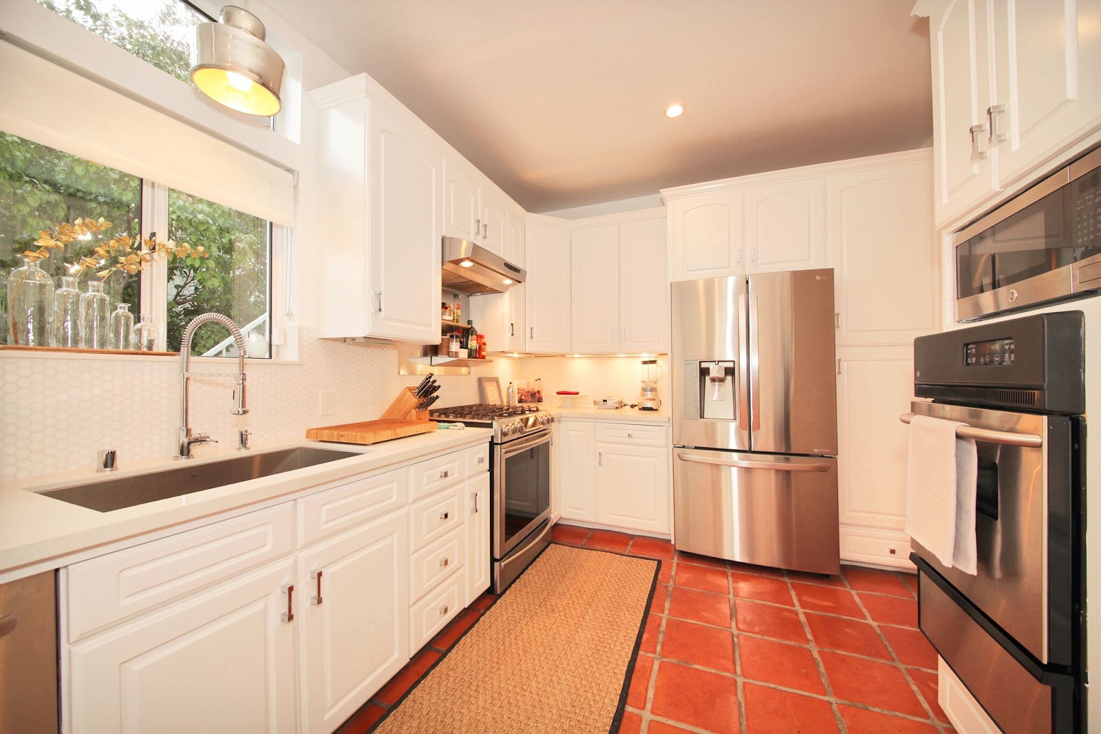 Interior modern kitchen with white cabinetry, stainless appliances, marble counters, terra cotta tile, and natural window light