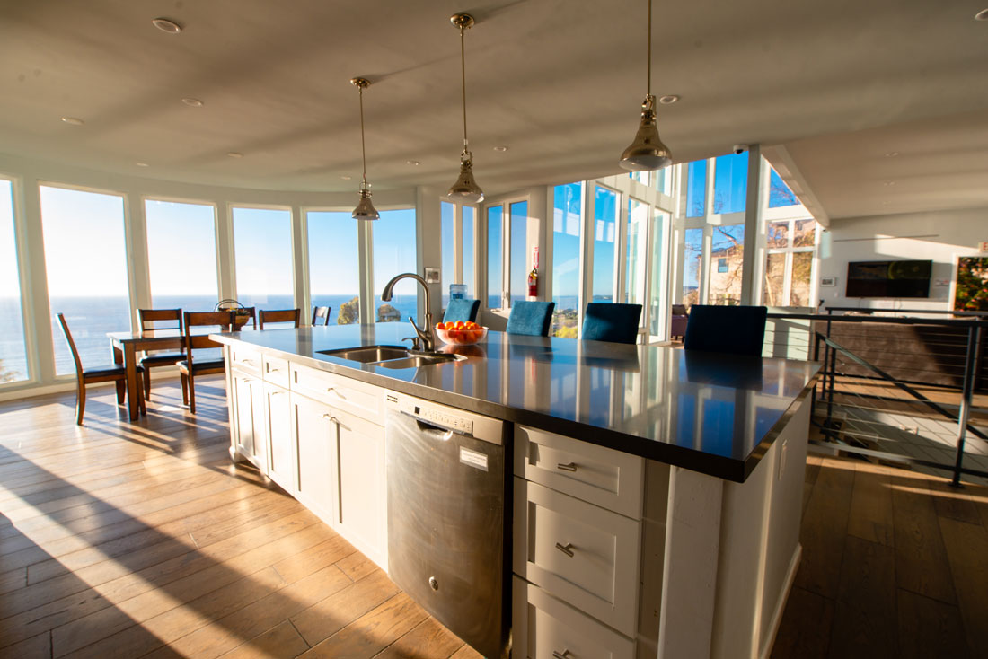 Interior kitchen featuring white cabinetry, dark countertop island, and large windows overlooking the ocean and pool area