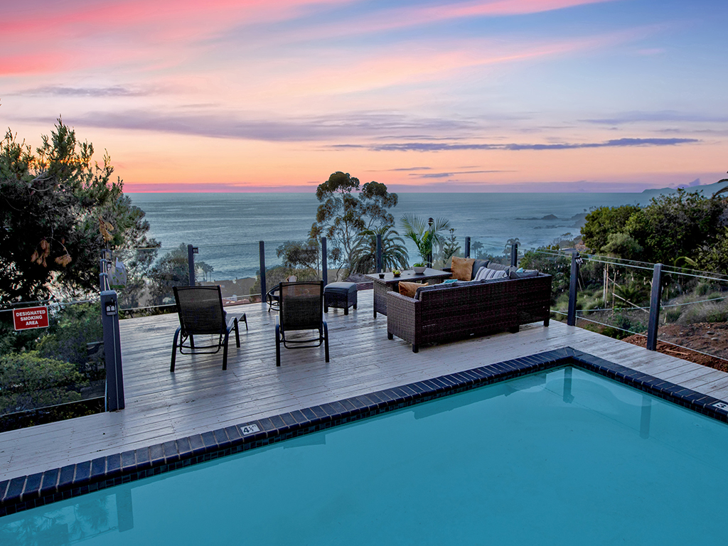 Outdoor pool deck with turquoise water, wooden decking, loungers, and expansive views of the Pacific Ocean horizon