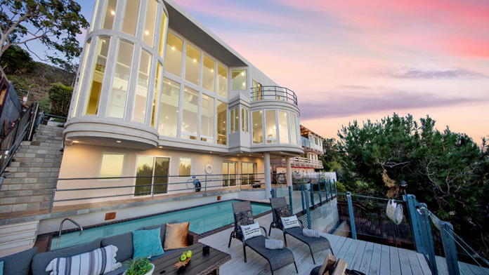 Exterior of modern building with curved glass walls, illuminated interior, heated pool, and ocean landscape at dusk