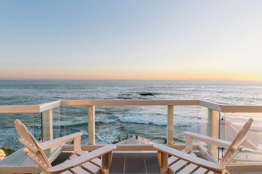 Exterior terrace with Adirondack-style chairs, modern glass railing, ocean and beach view at sunset or sunrise, peaceful seating area